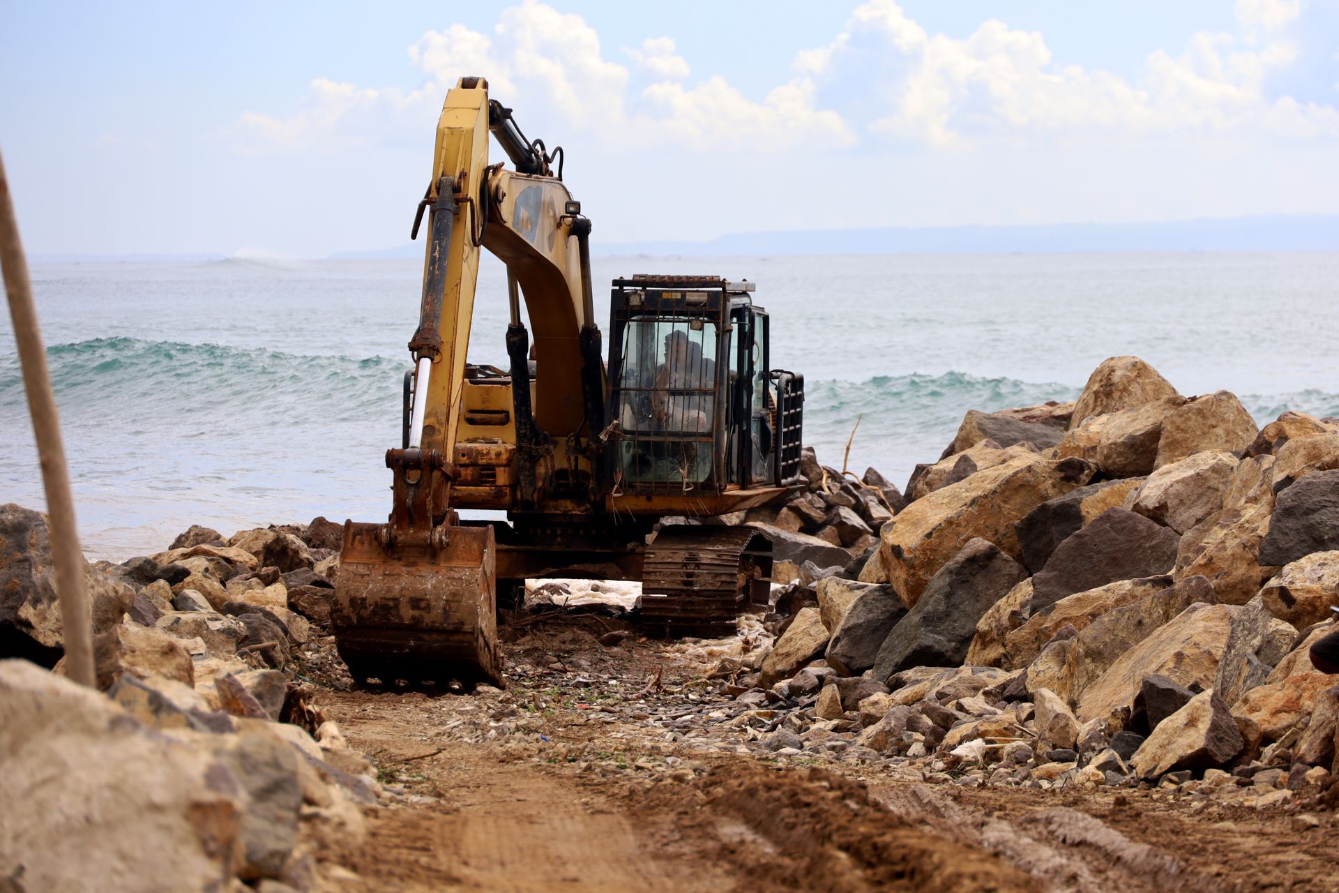 A yellow excavator is working on a rocky beach near the ocean.