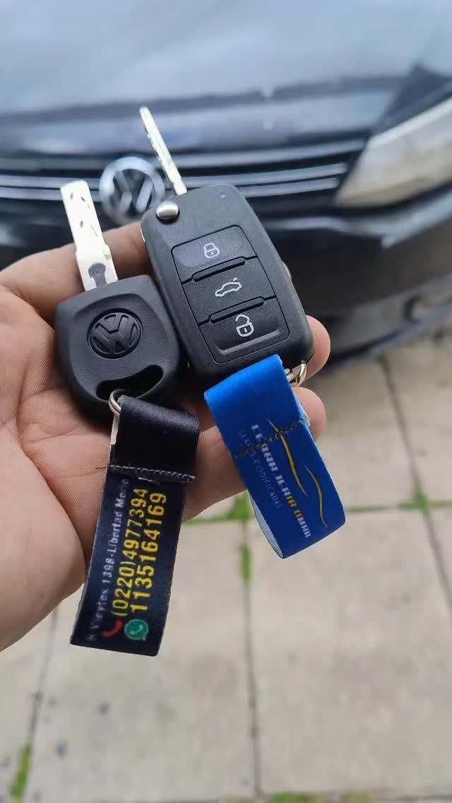 Mano sosteniendo las llaves de un coche Volkswagen con mandos y llaveros, delante de un coche.