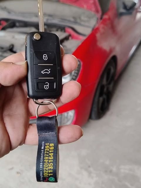 Una mano sostiene un llavero con la llave de un coche; al fondo se ve un coche rojo.