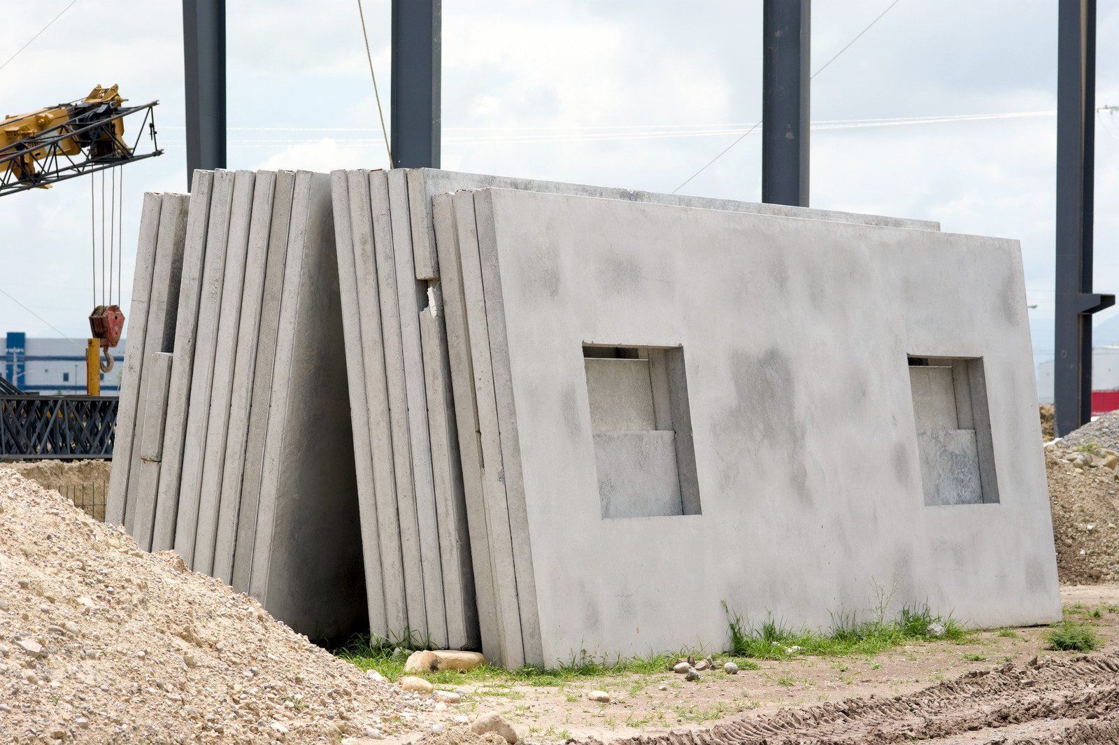 Concrete wall panels with window openings stacked outdoors near a construction site.