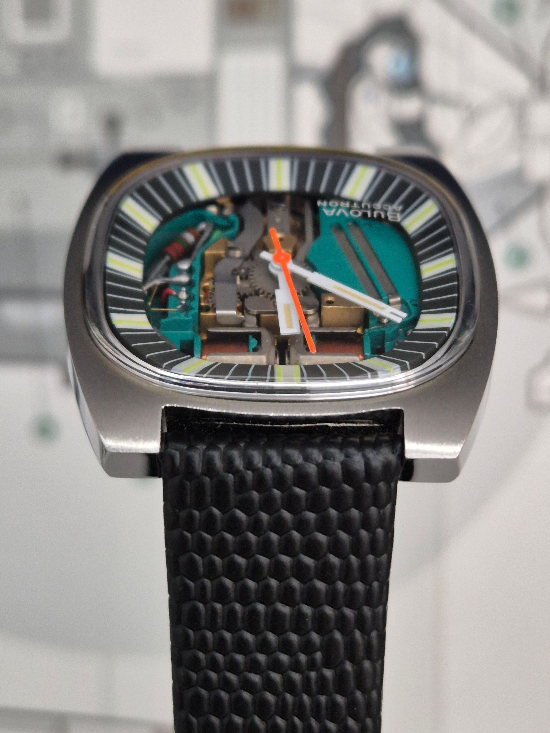 Silver wristwatch with skeleton dial, black strap, orange and white hands, set against a blurry backdrop.