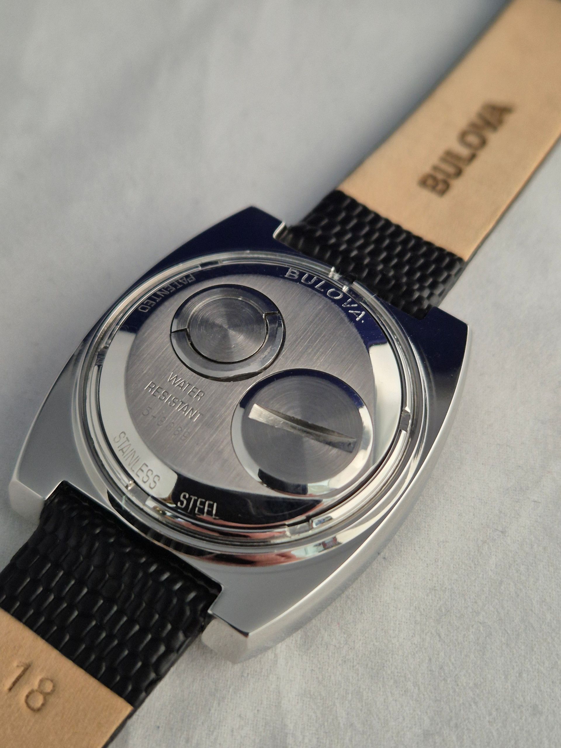 Back of a silver Bulova watch on a black strap. The watch has two circular components and a leather band.