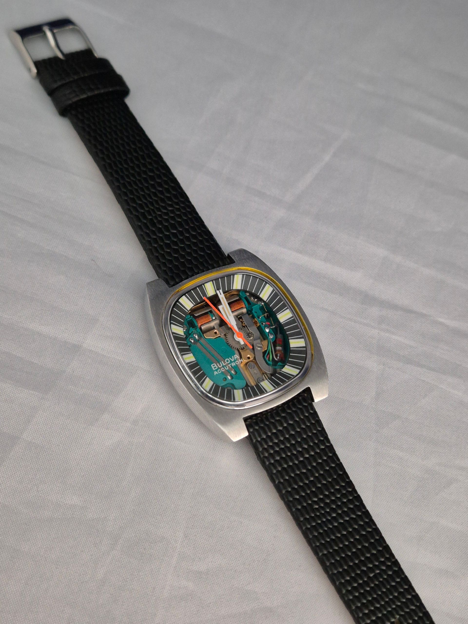 Silver wristwatch with a rectangular face, black strap, and visible green circuit board.