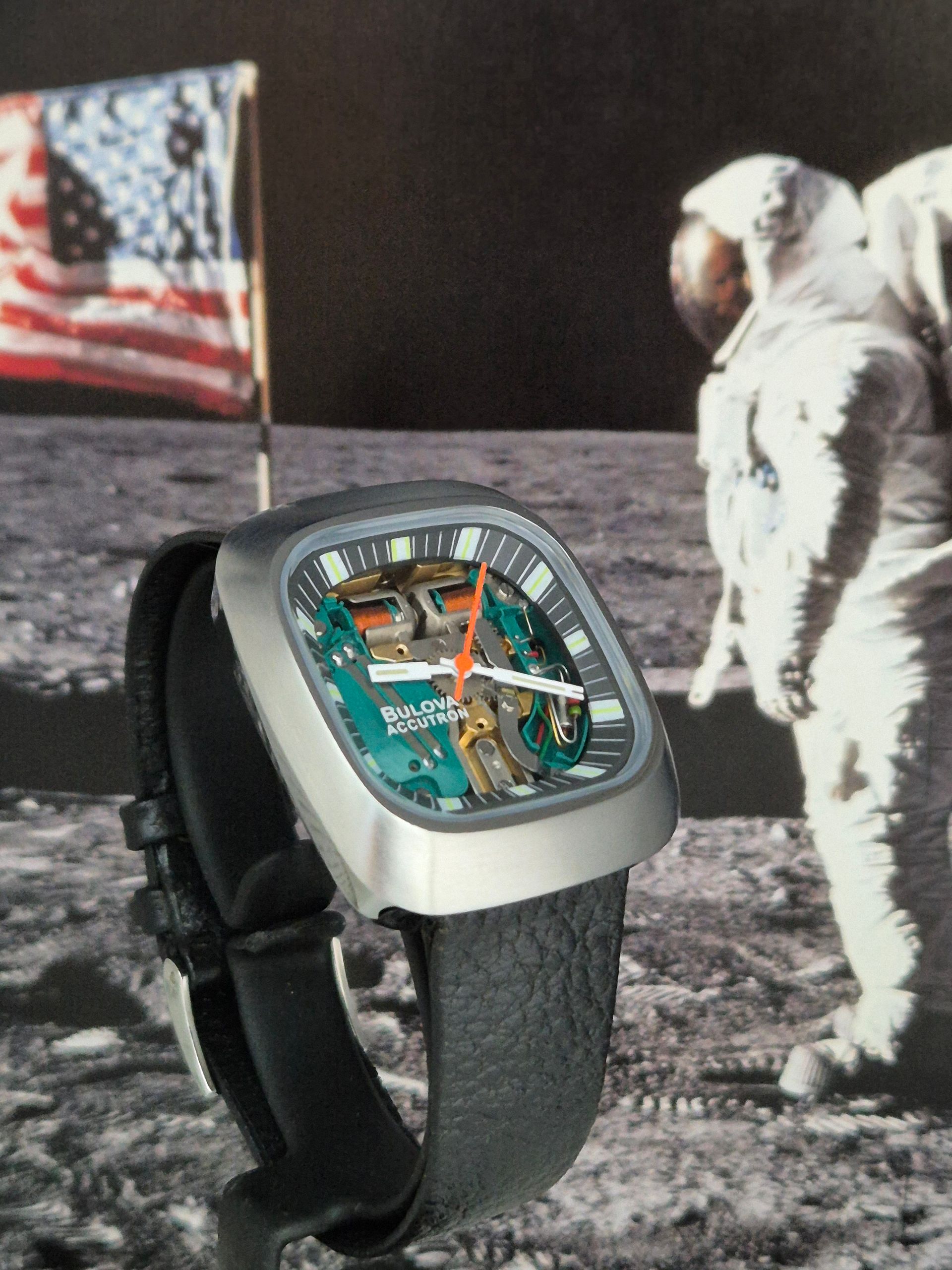 Square teal watch with black band in front of an astronaut on the moon with American flag.