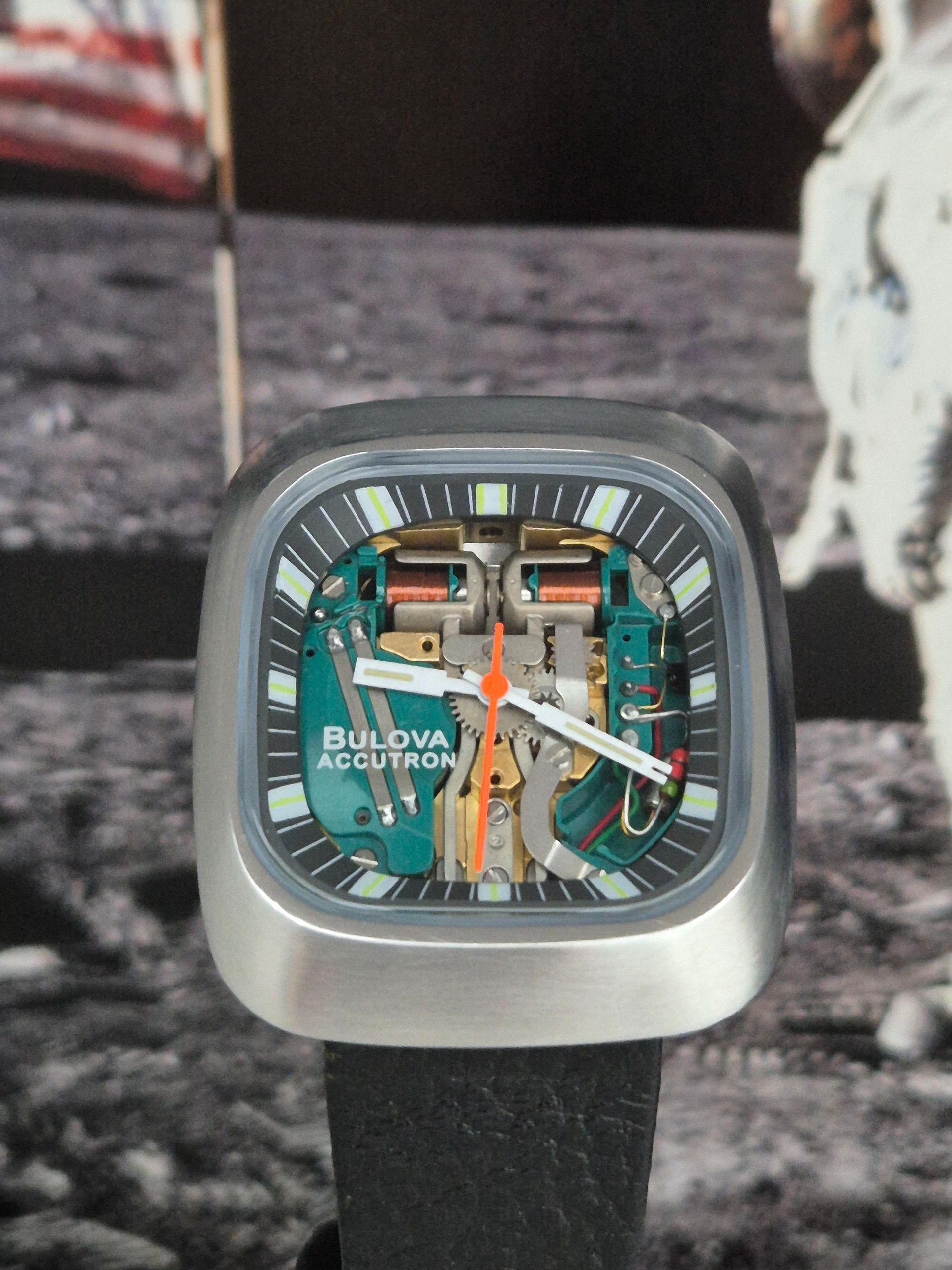 Bulova Accutron watch with exposed movement, silver case, black strap, against a lunar surface backdrop.