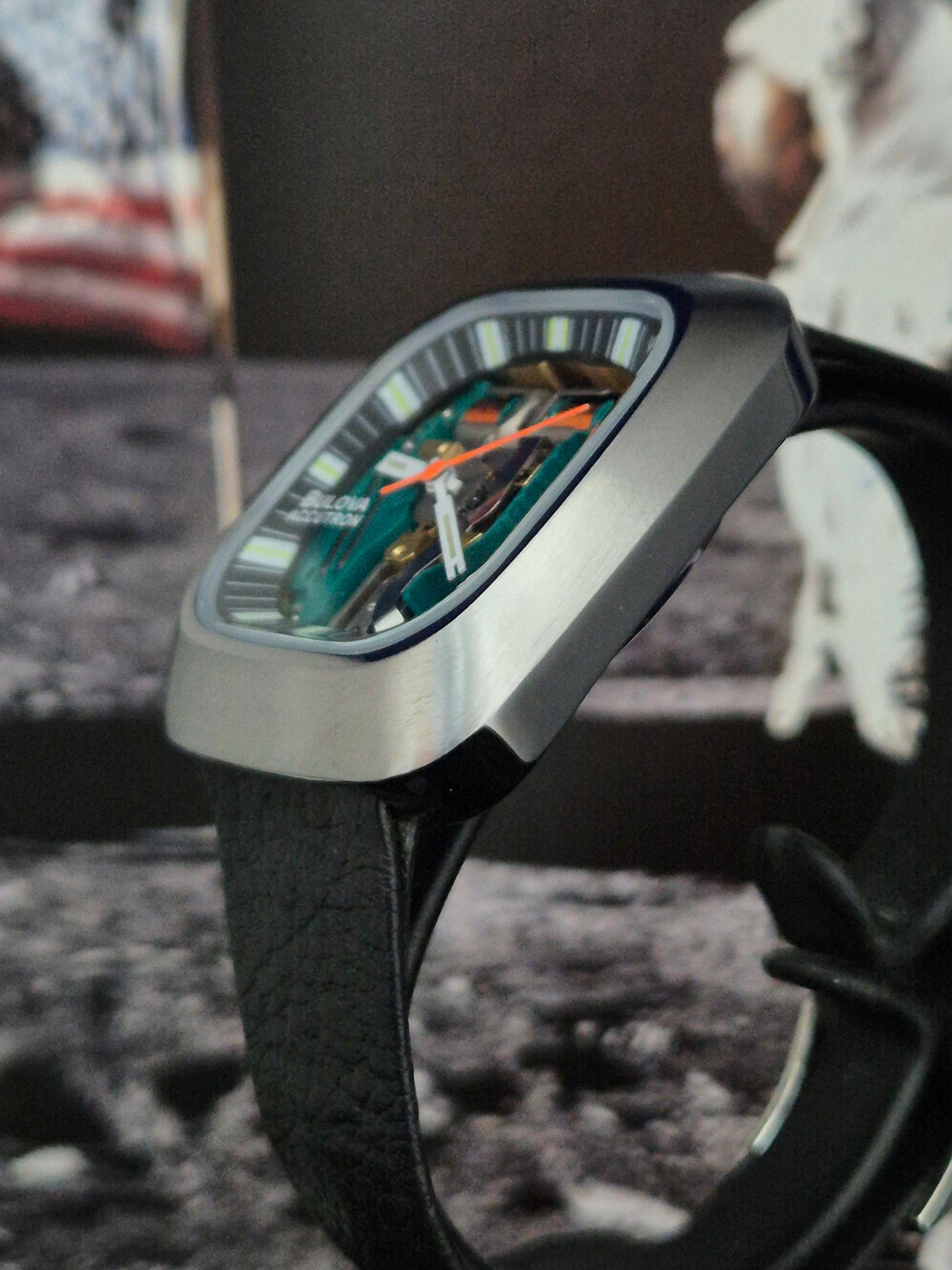 Silver wristwatch with green dial and orange accents; black leather strap; lunar backdrop.