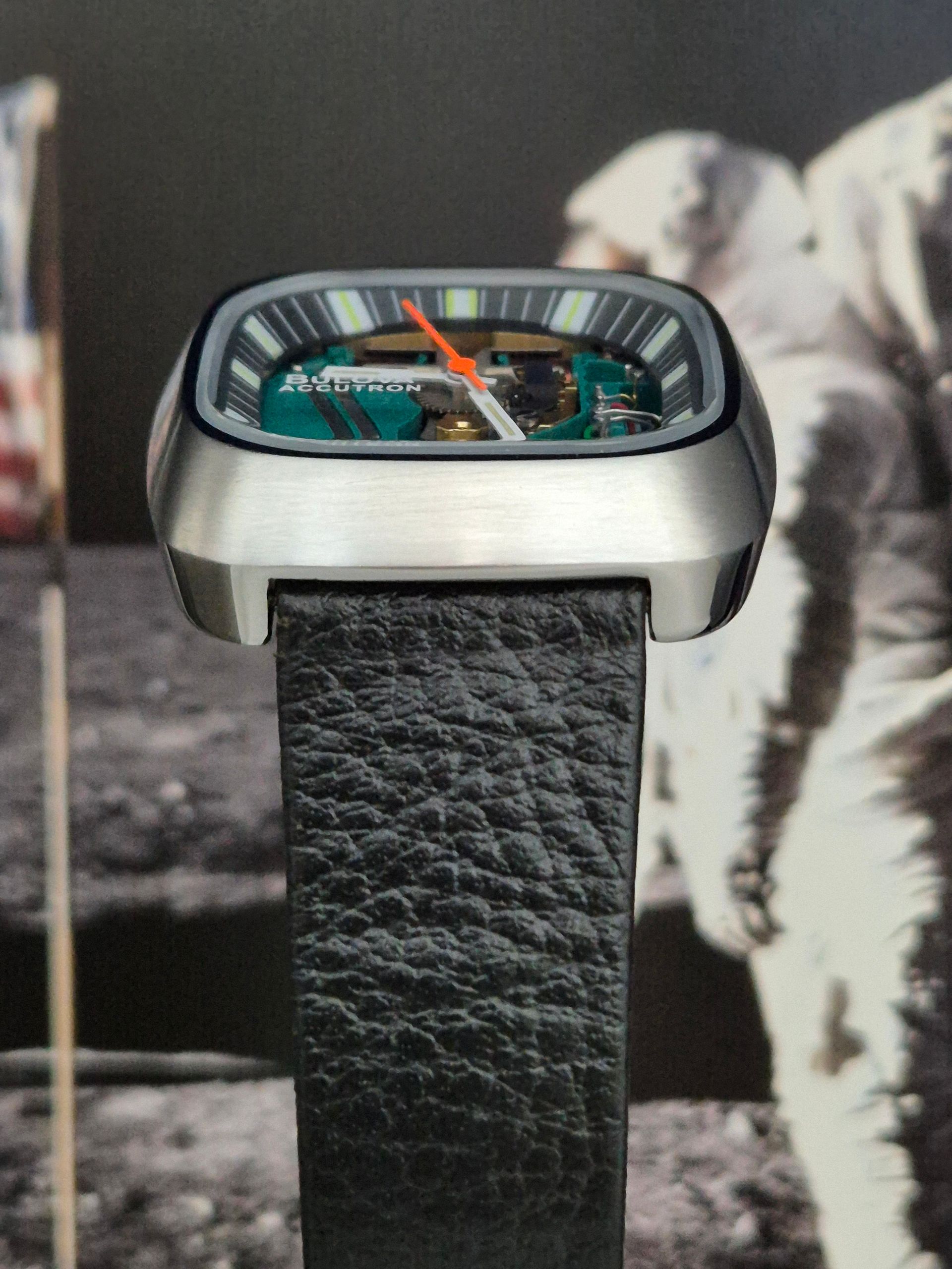 Square watch with green face, orange hand, and black leather band, against moon landing background.