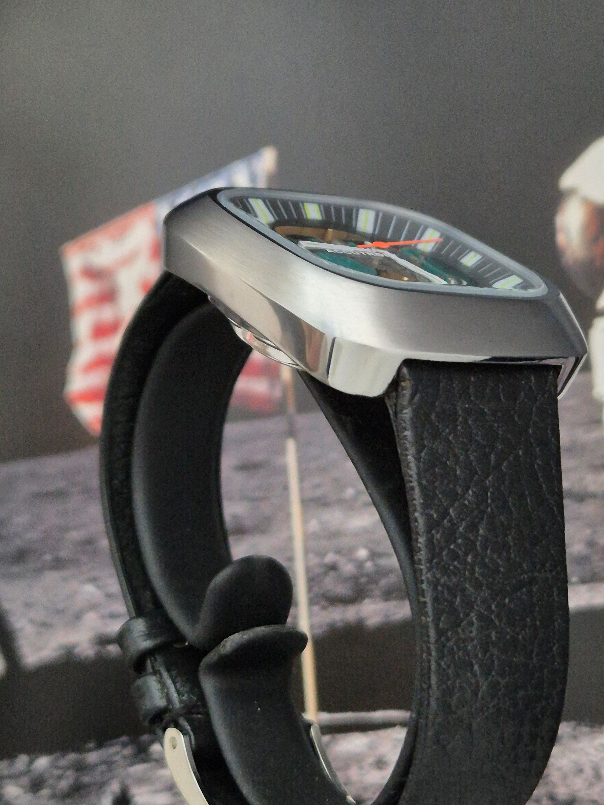 Silver wristwatch with black leather strap, green dial, and red hands, set against a lunar landscape and American flag.