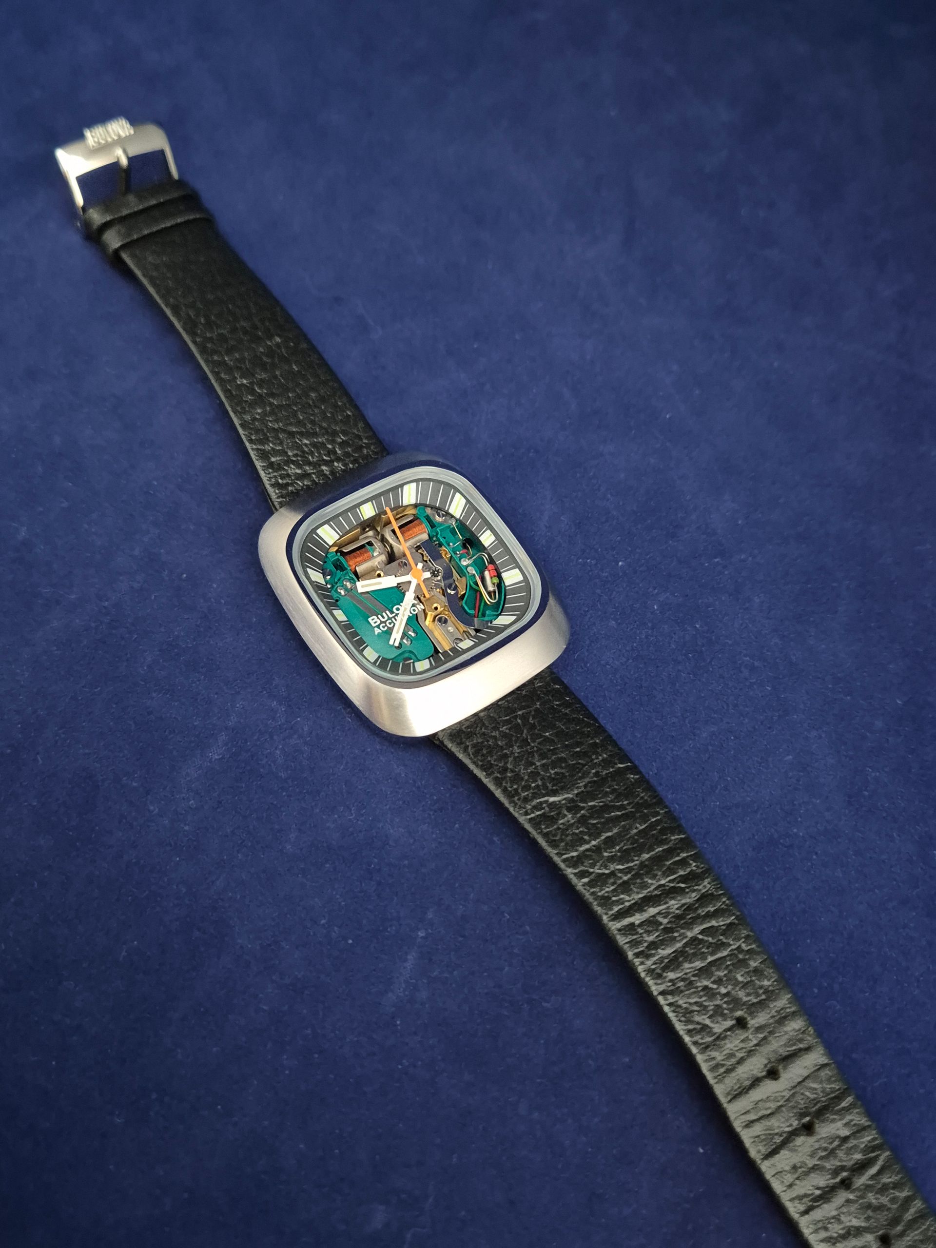 Silver wristwatch with a rectangular face, black band on blue background.