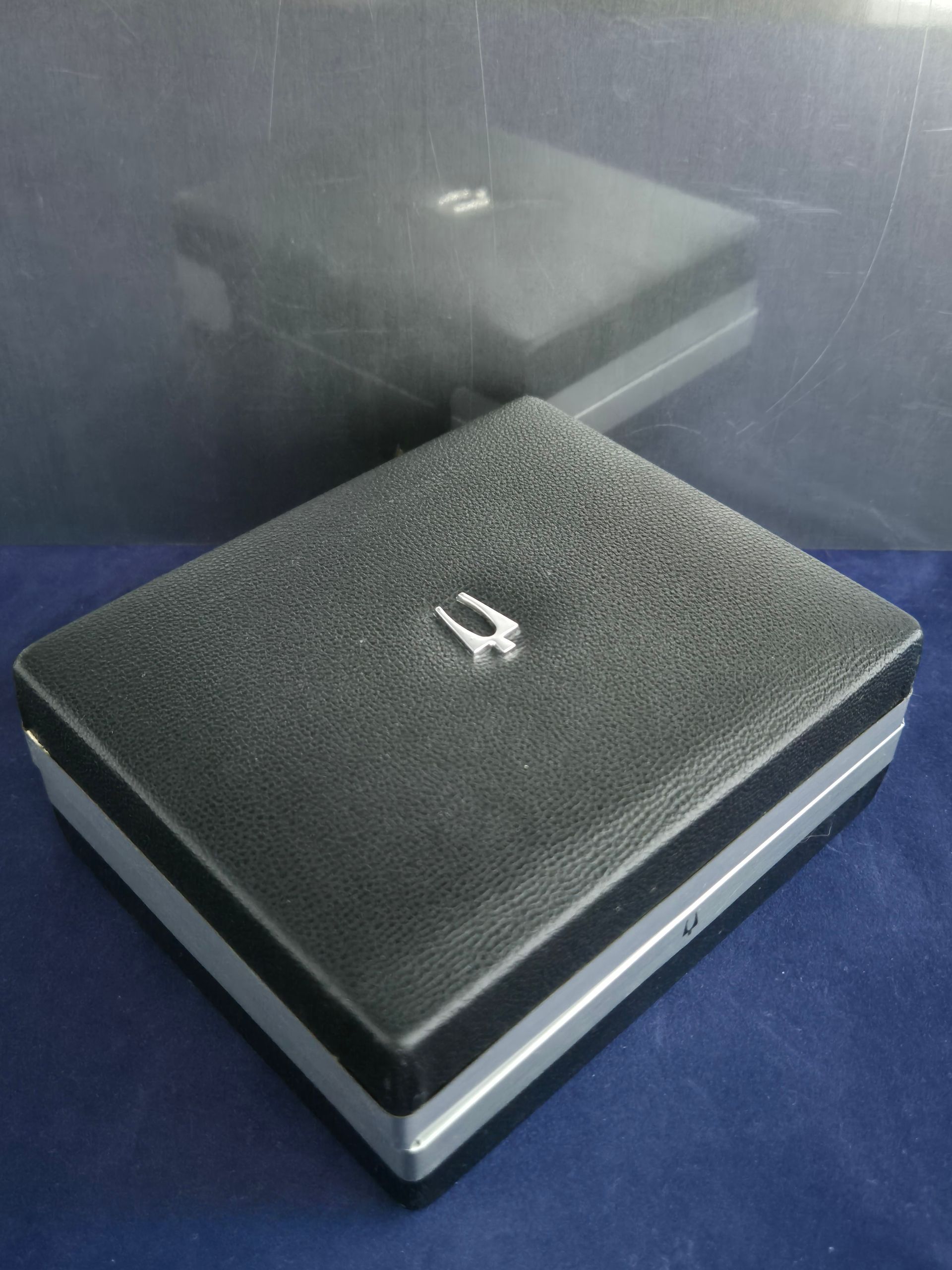 Black box with silver trim, small silver ornament on top. Reflection of box visible.