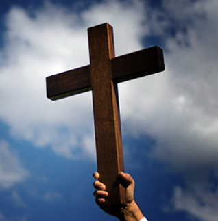 Hand holding a wooden cross against a blue sky with clouds.