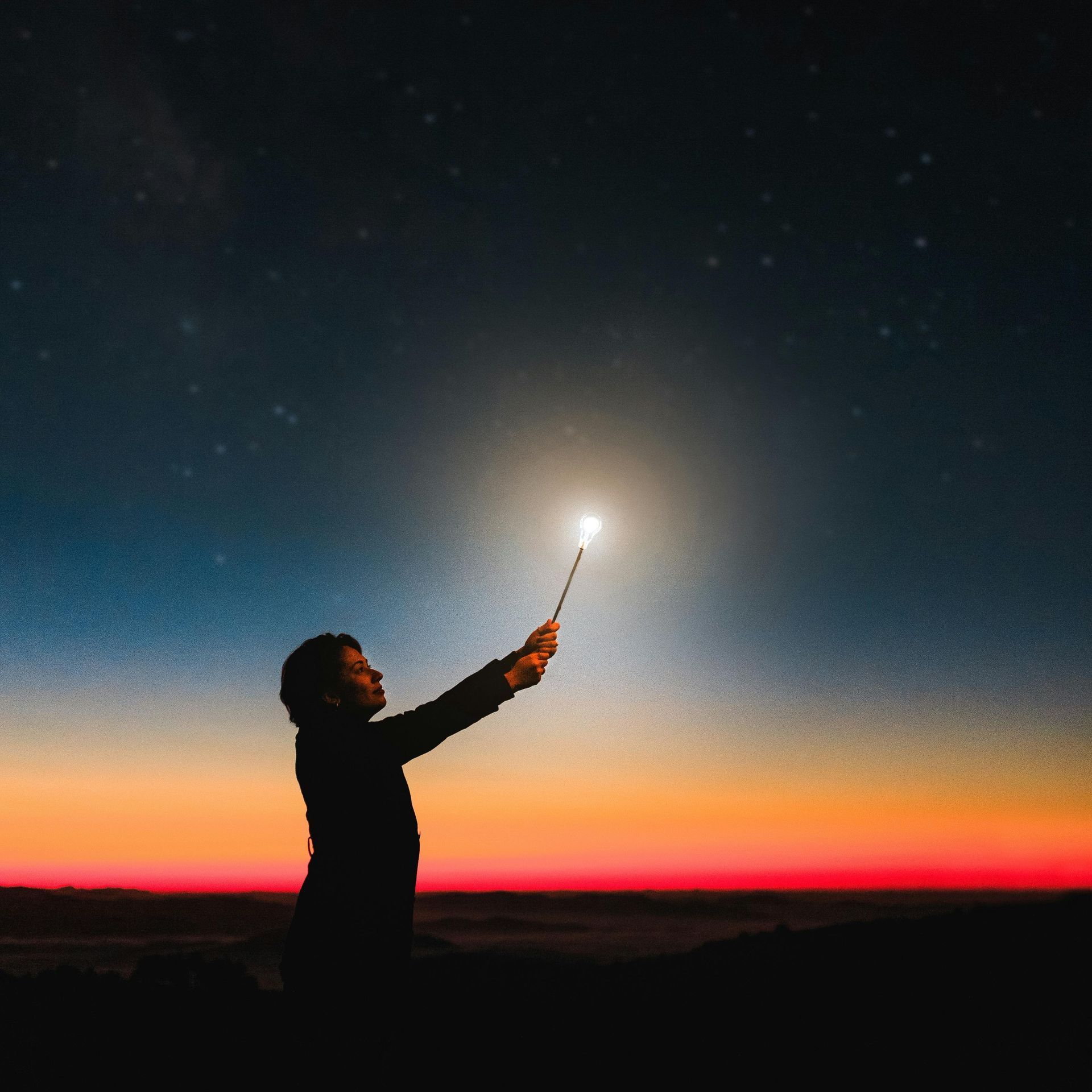 Silhouette of person holding sparkler against starry sky during a colorful sunset.