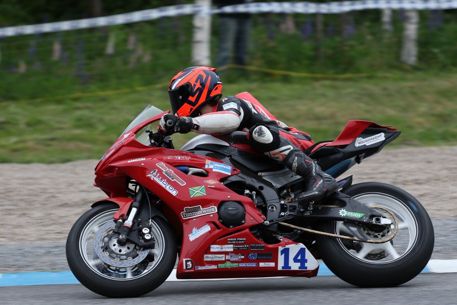 Motorcoureur leunt in een bocht op een rode motor, nummer 14, op een circuit. De rijder draagt een rood-zwarte helm en leren kleding.