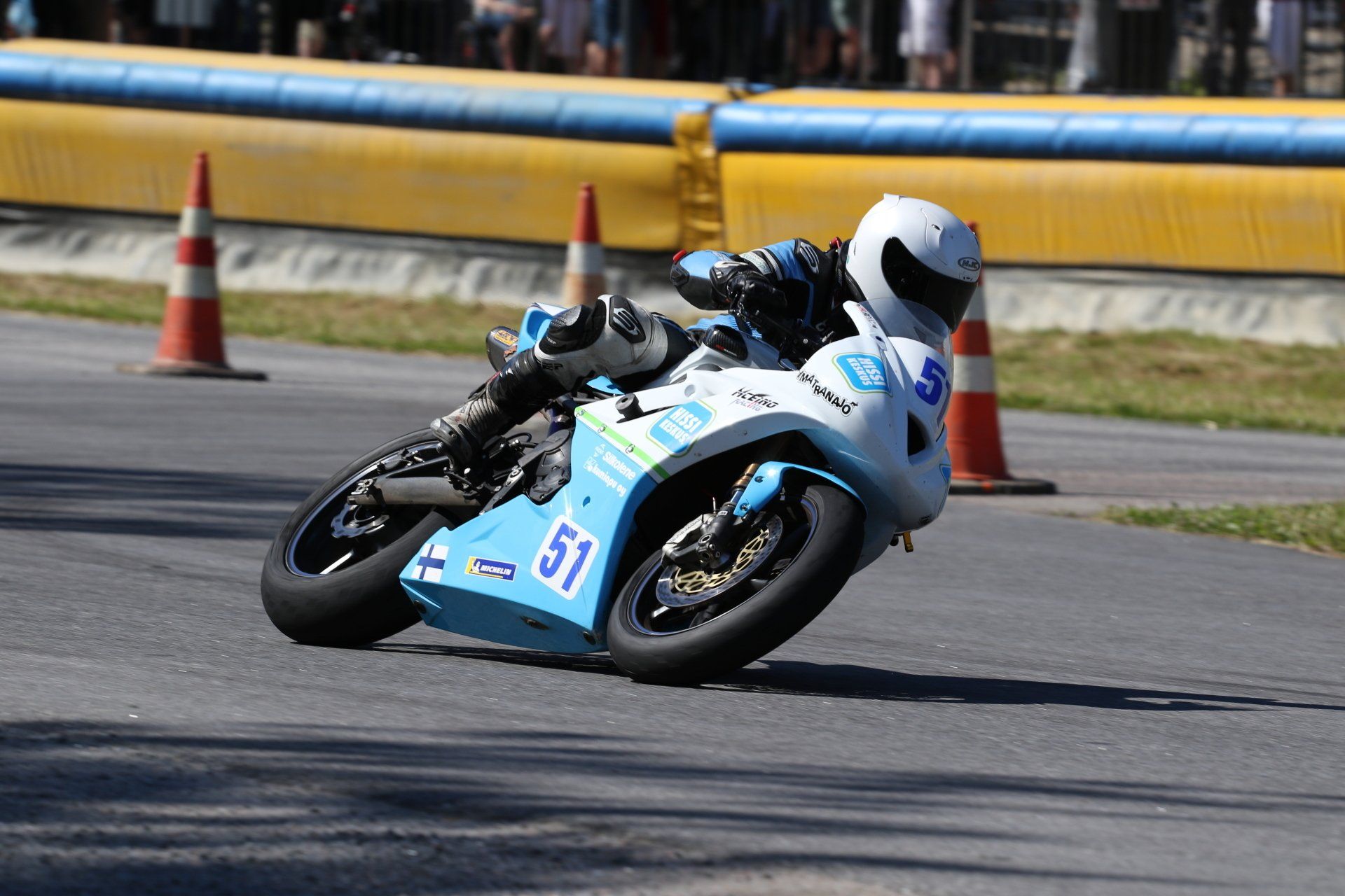 Motorcoureur met witte helm buigt zich voorover op een racecircuit. De motor is blauw en wit, oranje kegels op de achtergrond.
