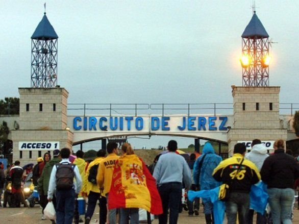 Mensen betreden Circuito de Jerez, een renbaan, onder een poort met blauwe letters. Twee torens flankeren de ingang.