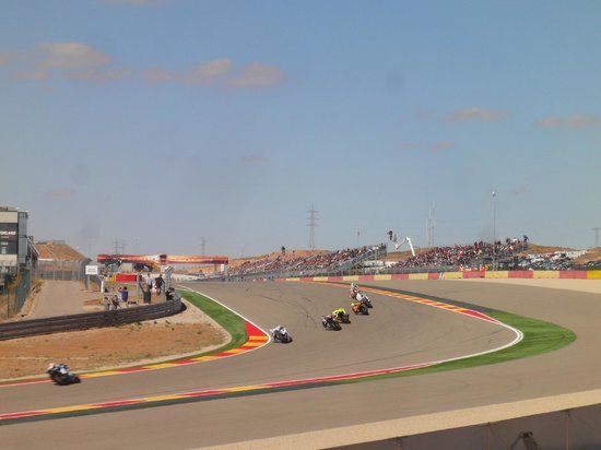 Motorrace op een bochtige baan, waarbij meerdere coureurs met hun rug naar de bocht staan. Op de achtergrond zijn toeschouwers te zien onder een blauwe lucht.