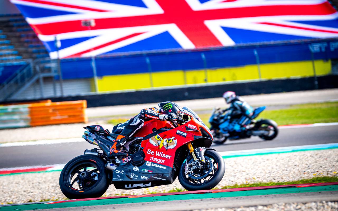 Twee motorrijders racen op een circuit, de een op een rode fiets die de bocht induikt, de ander op een blauwe fiets die erachteraan rijdt, met een Britse vlag op de achtergrond.