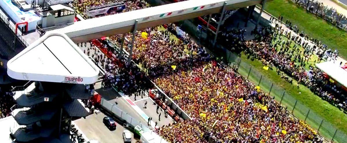 Luchtfoto van een Formule 1-circuit met een drukke tribune en een brug over de baan. Groen gras omzoomt de baan.