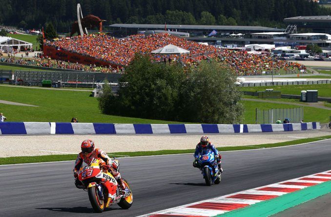 Twee motorrijders racen op een circuit, met een volle zaal op de achtergrond. De setting is een groen, zonnig racecircuit.