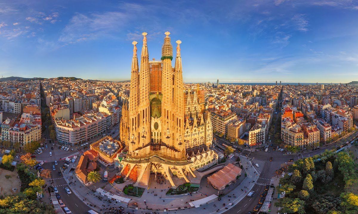 De Sagrada Familia in Barcelona, Spanje, omringd door stadsgebouwen onder een helderblauwe hemel. De stenen gevel van de kerk wordt verlicht door zonlicht.