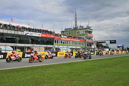 Motoren staan opgesteld aan de startgrid van een race, klaar om te beginnen. Op de achtergrond een racecircuit met tribunes en een verkeerstoren.