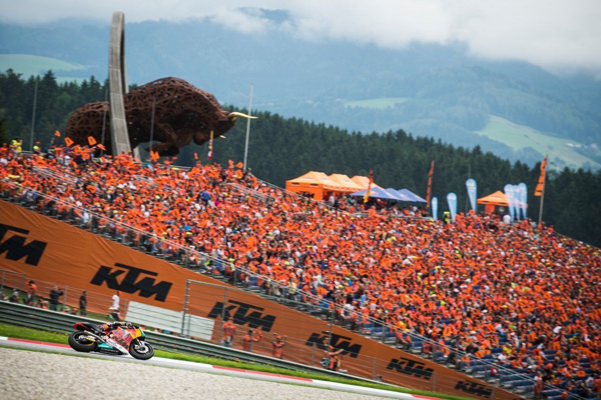 Een motorrace op een circuit met oranje fans op de tribune. Bergen en bomen vormen de achtergrond.