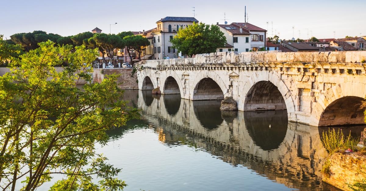Stenen brug met bogen over een kalme rivier. Gebouwen langs de rivieroever en bomen omlijsten de voorgrond.