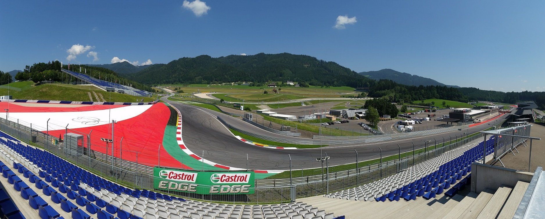 Panoramisch uitzicht op een renbaan met rood-witte tribunes, een kronkelend asfaltparcours en groene bergen op de achtergrond onder een blauwe lucht.