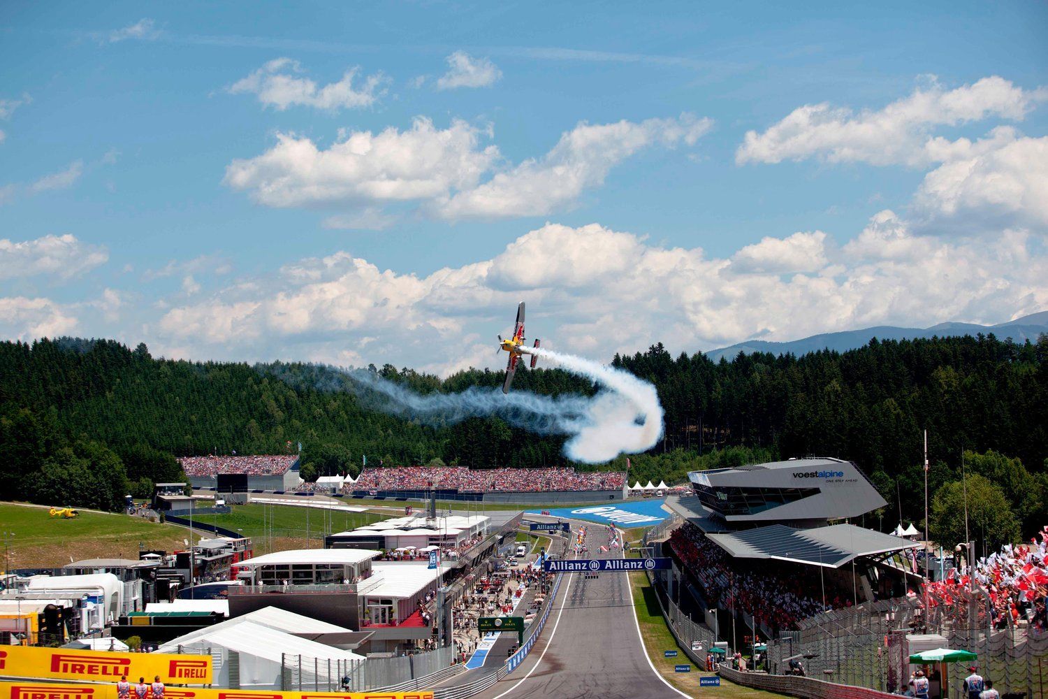 Een vliegtuig dat acrobatische toeren uithaalt boven een Formule 1-circuit. Witte rooksporen tegen een blauwe lucht en groene bergen.