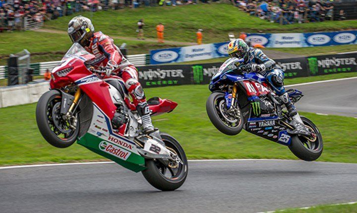 Twee motoren maken wheelies op een circuit. De rood-witte Honda loopt voor op de blauw-zwarte Yamaha, beide met verhoogde voorwielen.