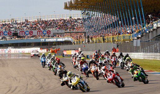 Motorrace op een circuit met toeschouwers op de tribune. Een groep rijders leunt in een bocht.