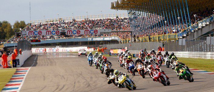 Motoren racen op een circuit voor een vol stadion. Motorrijders leunen in een bocht, toeschouwers kijken toe vanaf de tribune.