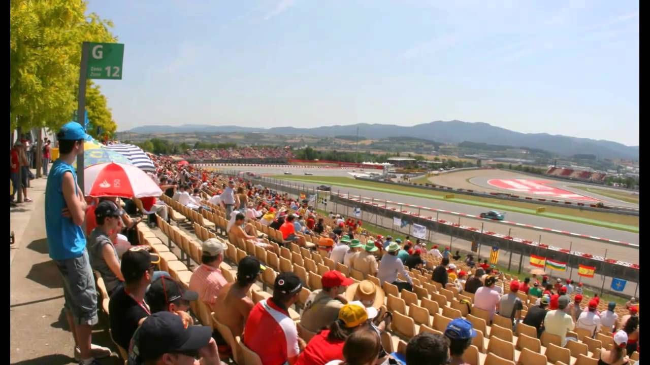 Toeschouwers kijken naar een race op een zonnige buitenrenbaan. Een blauwe raceauto raast langs de tribune vol mensen, met bergen op de achtergrond.
