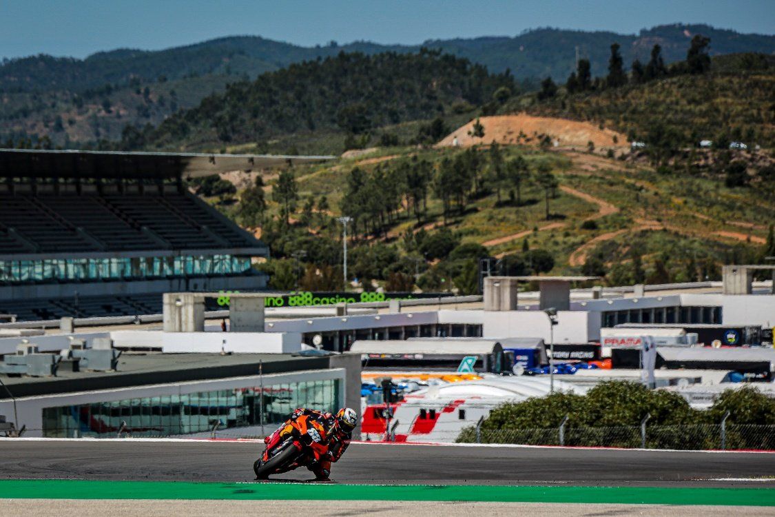Een motorcoureur leunt in een bocht op een circuit, met gebouwen, tribunes en heuvels op de achtergrond en een zonnige hemel.