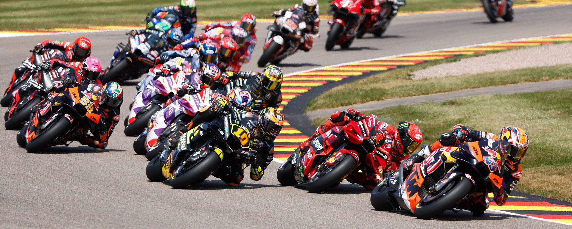 Motorrace op een circuit. Coureurs leunen in een bocht, motoren in beweging, en sommige zijn oranje, rood en zwart.