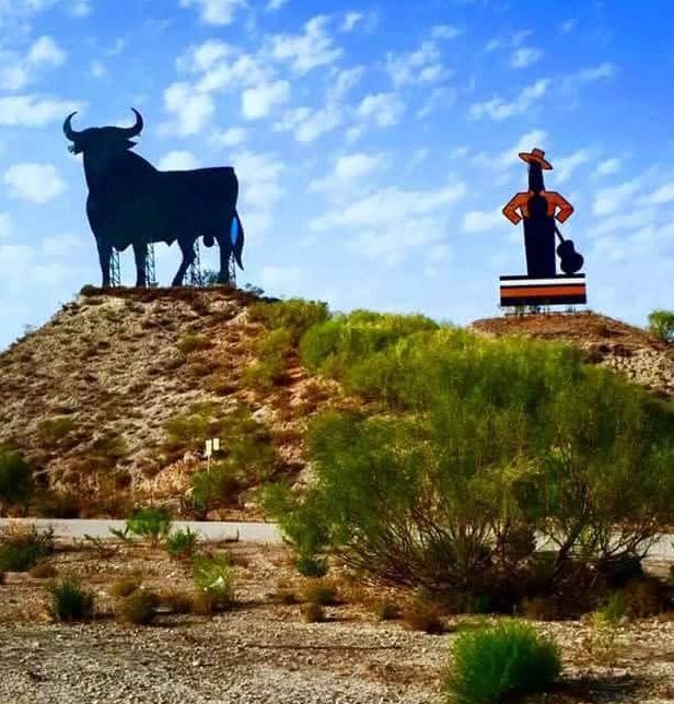 Silhouet van een zwarte stier en een flamencodanseres op heuvels tegen een blauwe lucht. Spaanse herkenningspunten langs de weg.