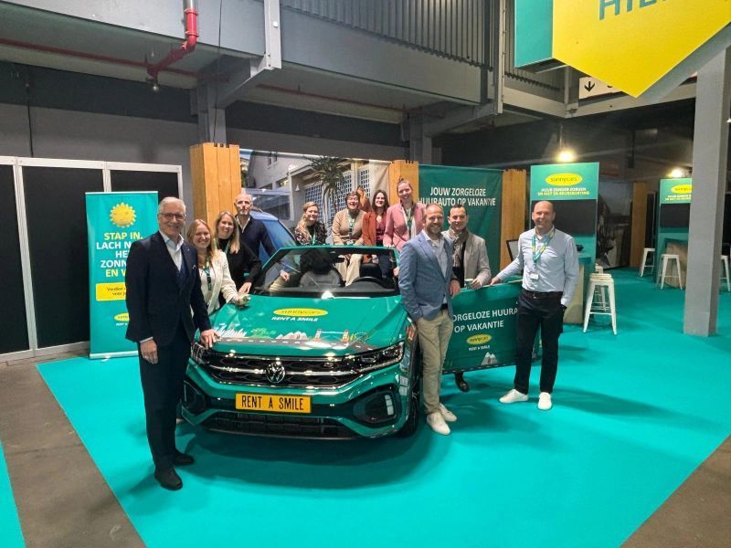 Een groep mensen rond een groene auto op een beursstand met het logo van het bedrijf erop.