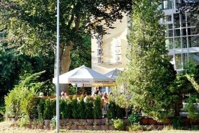 Terras met witte parasols en eethuisjes, naast een gebouw met een uithangbord waarop 'Hotel' staat. Groene bomen en struiken omlijsten het tafereel.