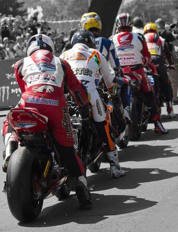 Motorcoureurs stonden aan de startlijn opgesteld, gekleed in kleurrijke racepakken en helmen, met op de achtergrond een menigte.