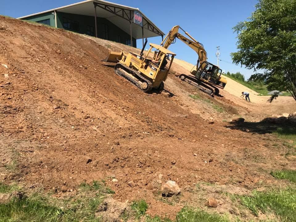 Two excavators working on a steep dirt slope beside a metal-roofed building under a blue sky