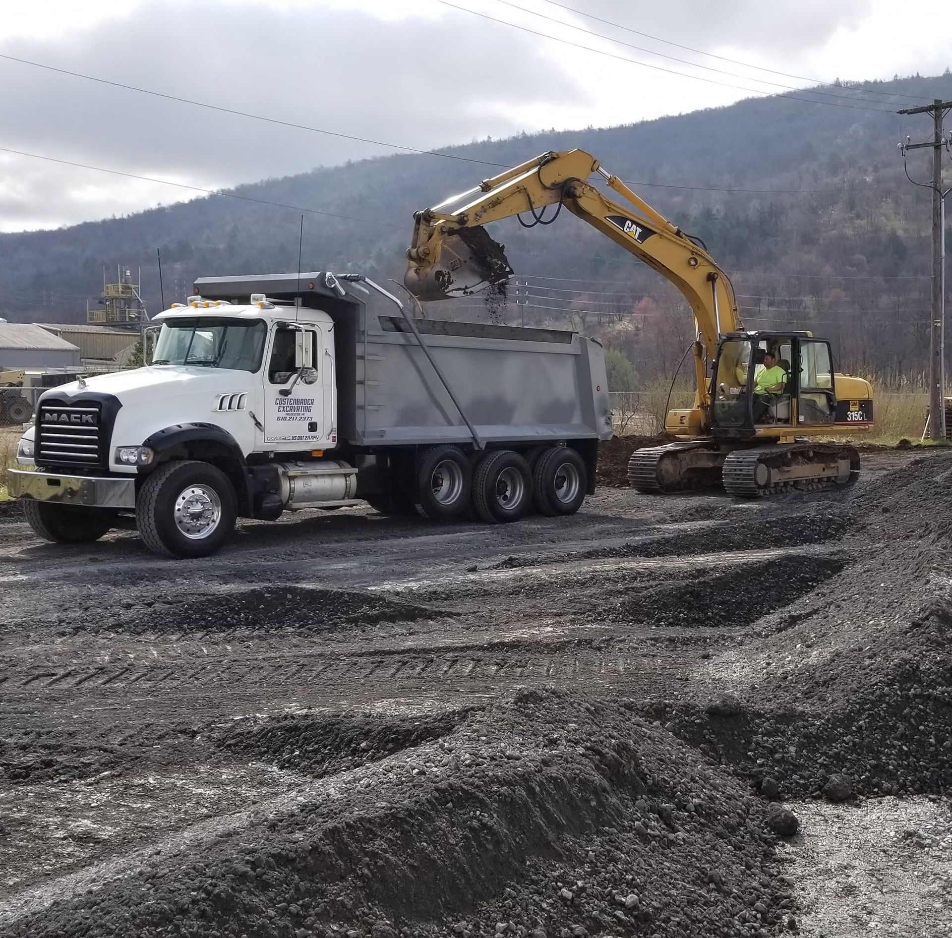 Excavator loads gravel into a white dump truck at a construction site with mountains in the background