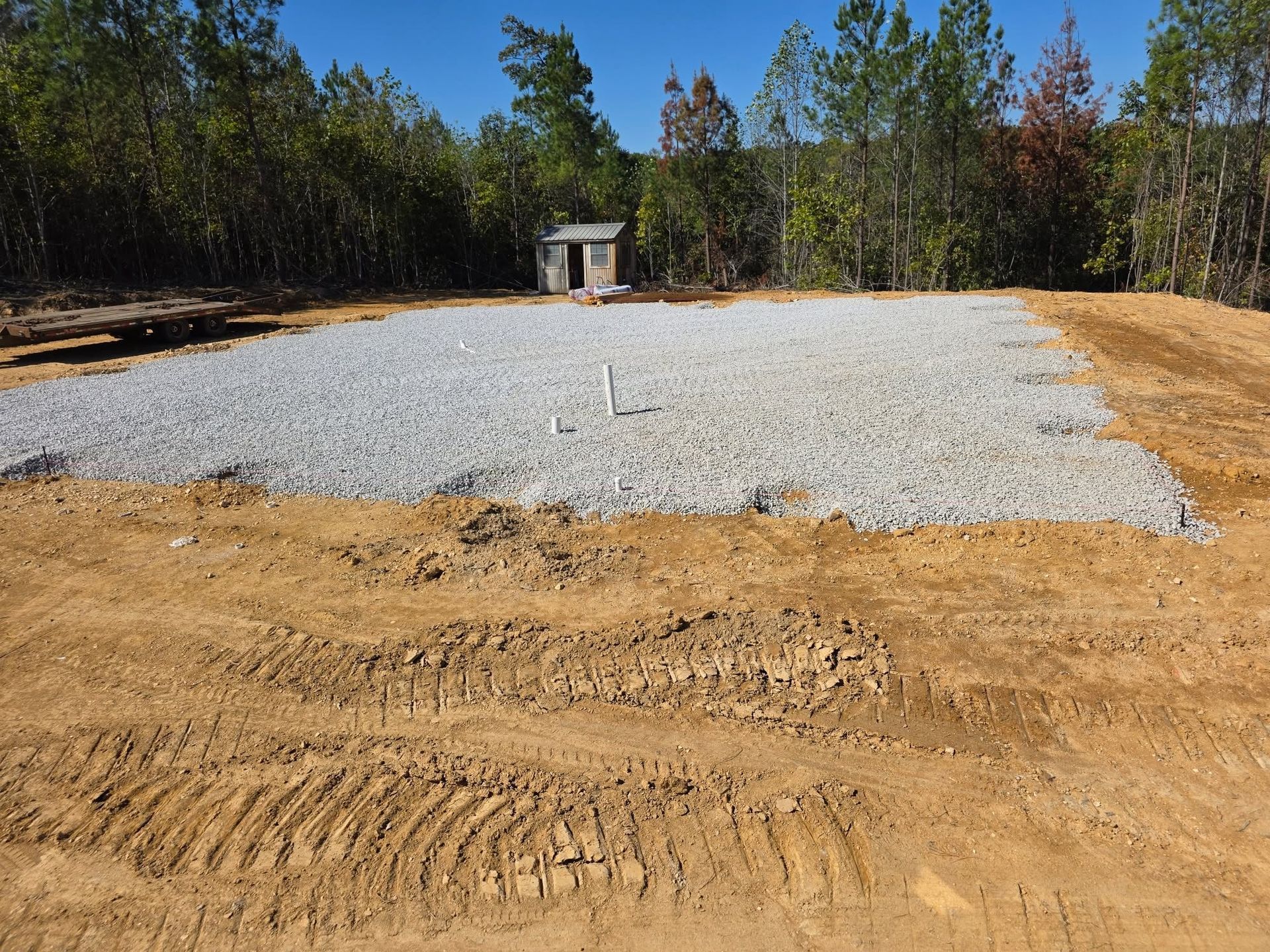 Gravel pad in a wooded clearing with a small shed and dirt foreground under a clear sky