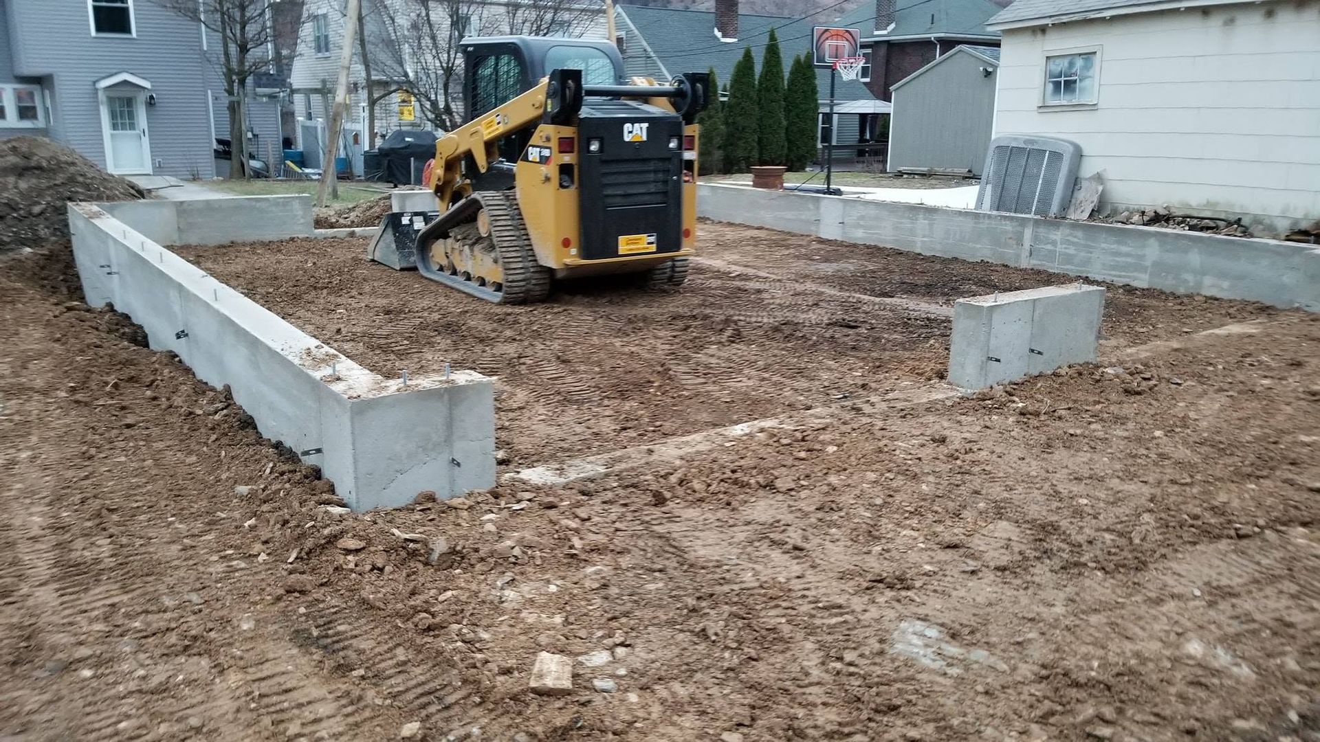 Mini excavator grading soil inside a concrete foundation site in a residential yard.
