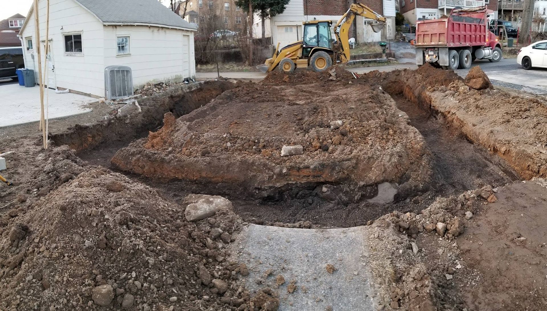 Construction site with muddy trench and excavator beside a road and houses