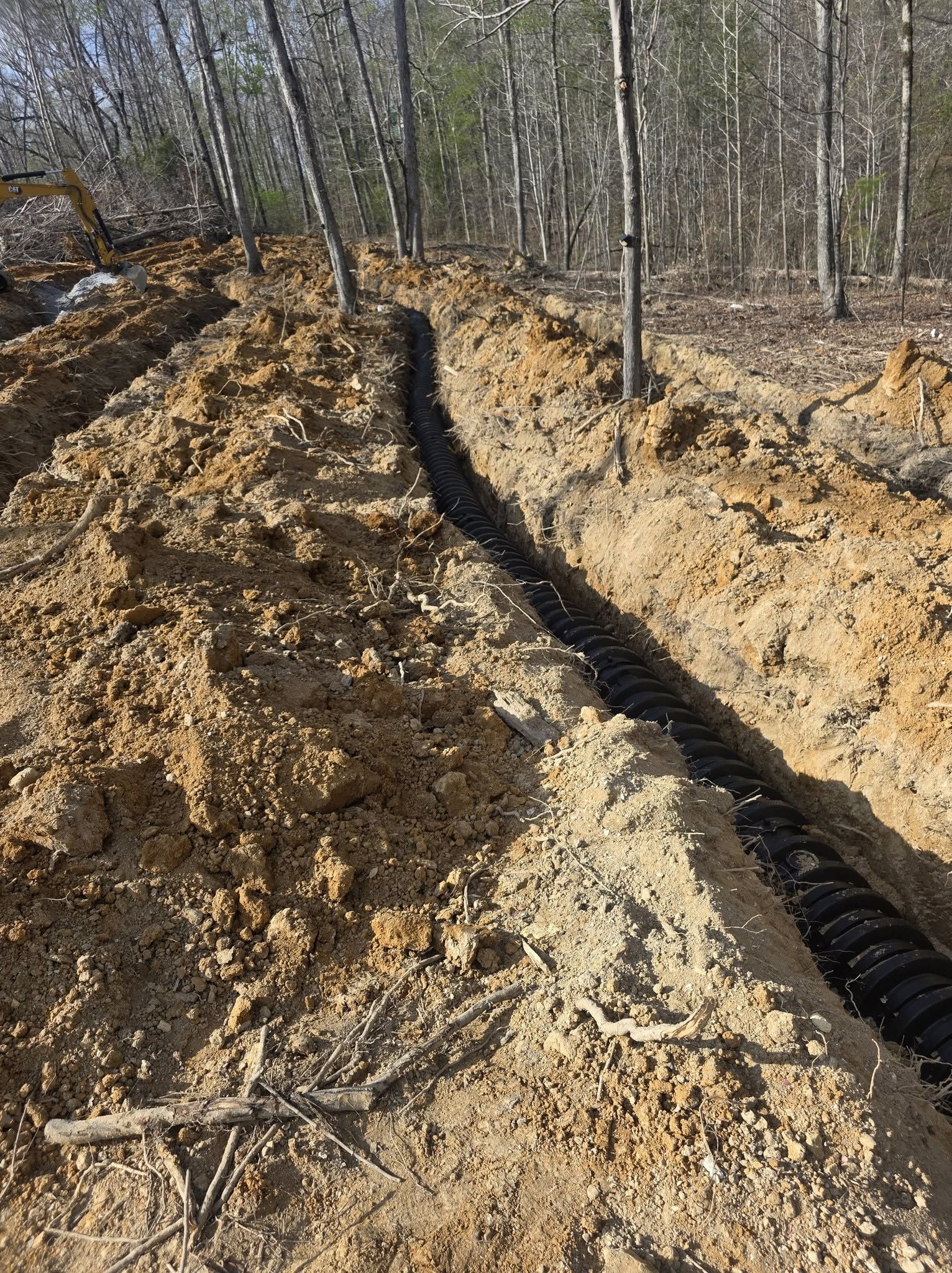 Dirt excavation trench in a wooded area with a black drainage pipe running along the right side.