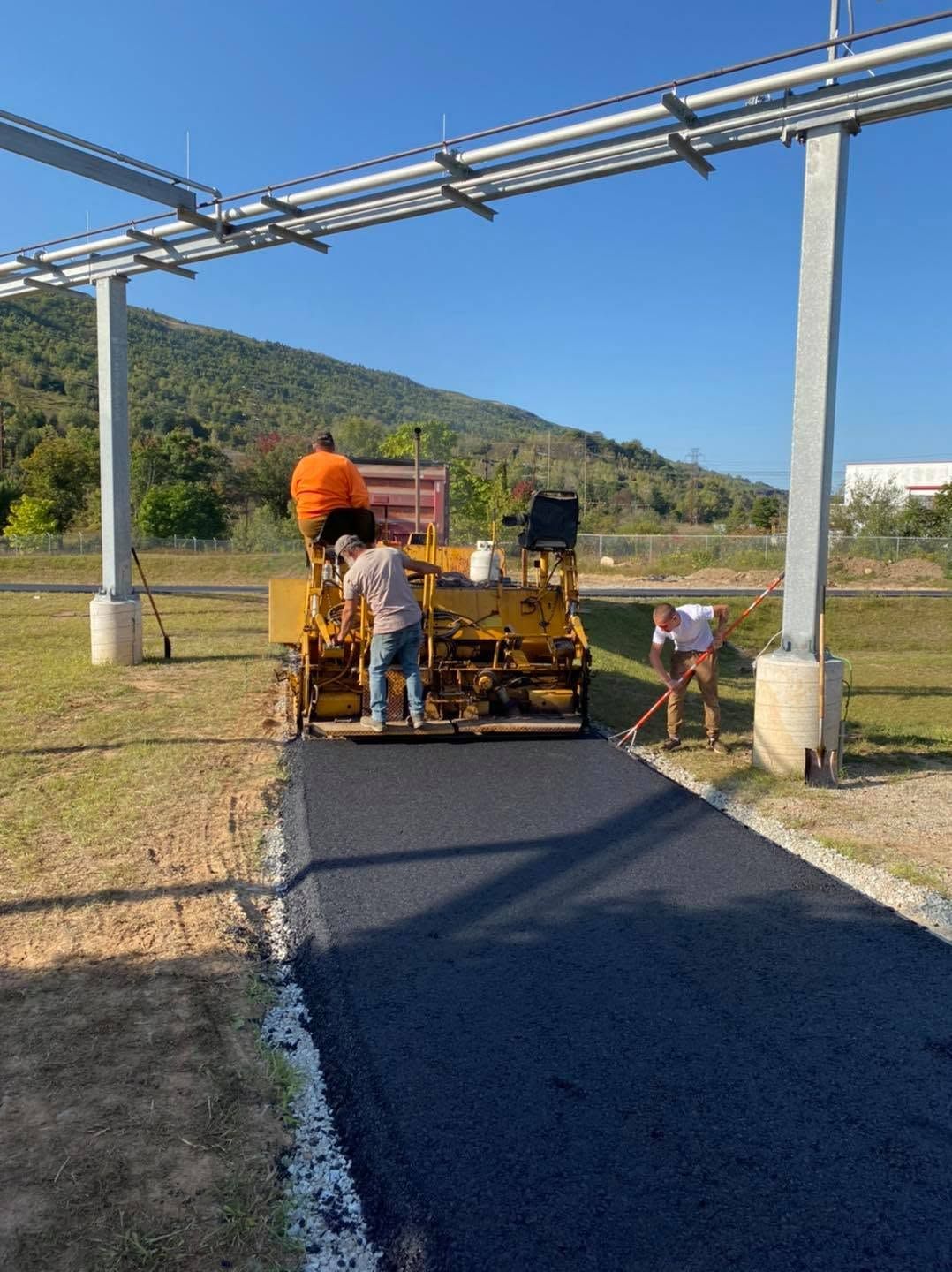 Workers paving a black asphalt road beside a metal structure in a rural field