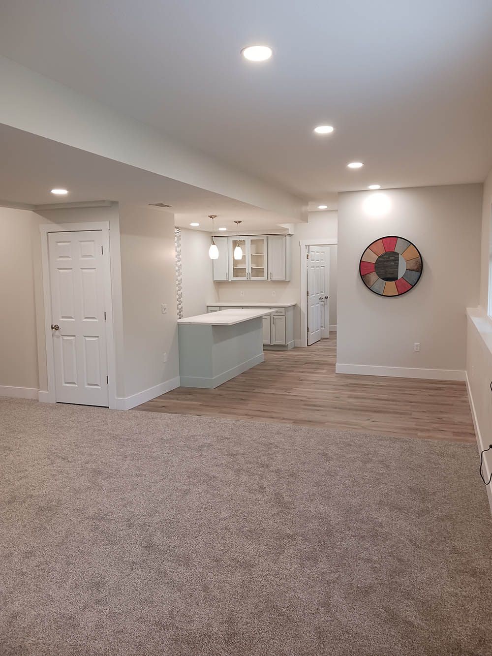 A basement with a carpeted floor and a kitchen in the background.