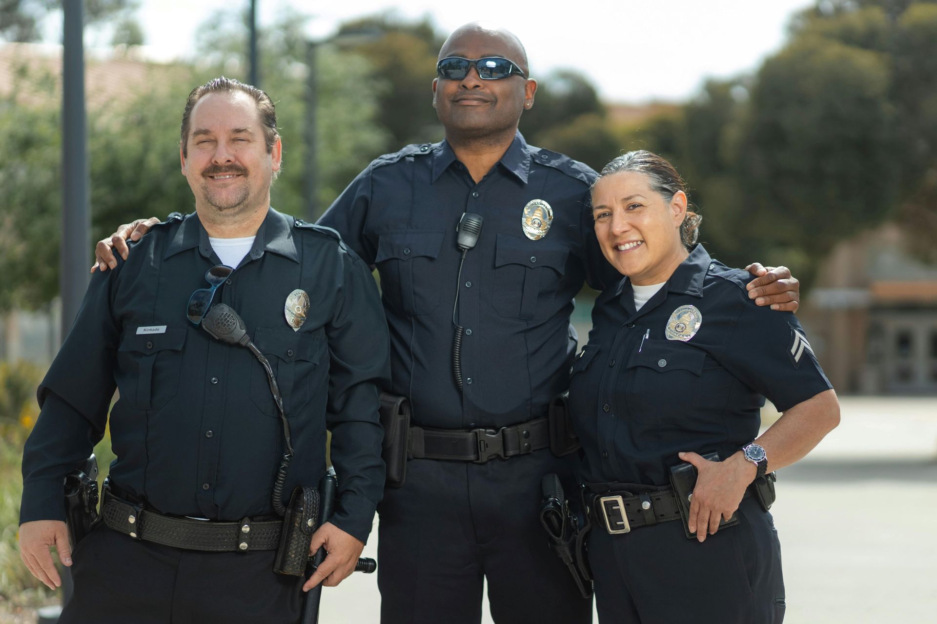 Three uniformed police officers stand side-by-side, smiling outdoors in a bright, natural setting.