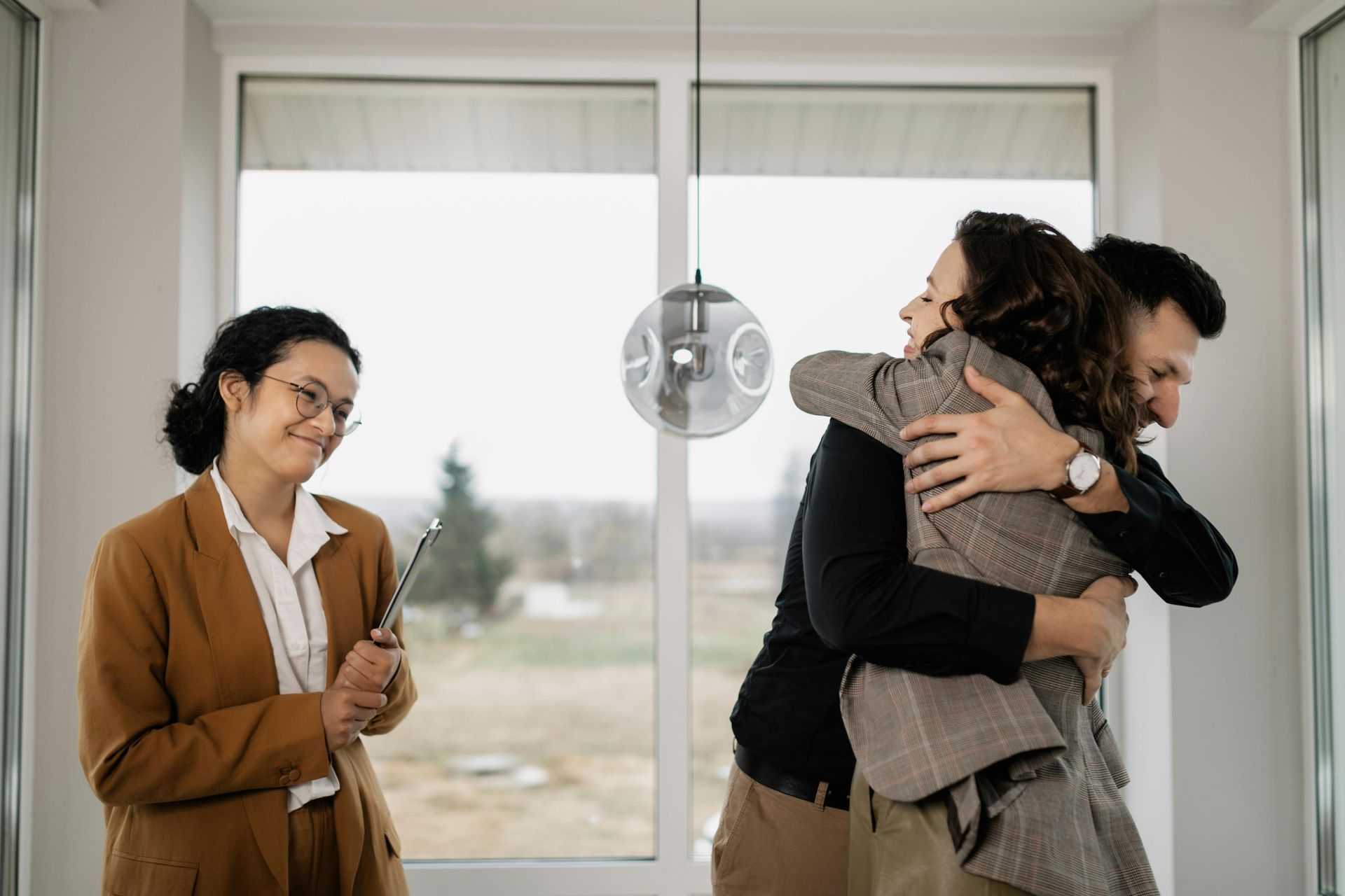 Three people by a window; two hug tightly while a woman smiles nearby.