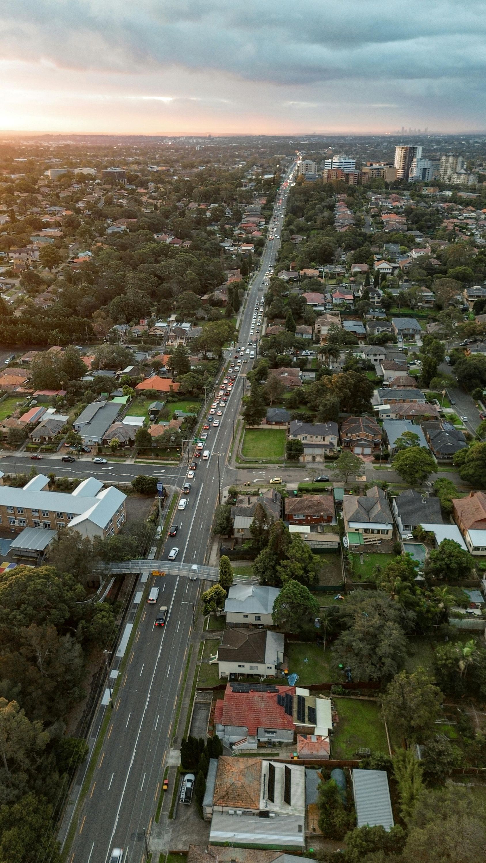 Aerial view of a city street lined with houses and trees under a cloudy sunset sky.